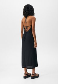Femme aux cheveux bouclés et foncés portant une robe midi noire dos nu et des sandales compensées noires, debout devant un fond clair uni.