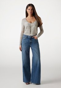 Cardigan tricotat de culoare gri deschis, cu modele texturate, purtat peste un top alb. Blugi blue cu talie înaltă și pantaloni largi. Sandale negre simple completează ținuta.