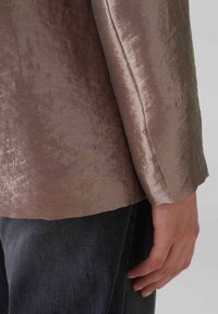 Hellbrauner Stoffblazer mit einem dezenten Glanz, im geraden Schnitt und mit taillierten Ärmeln, kombiniert mit dunkler Jeans.