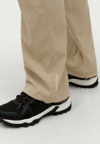 Beige buitenbroek met een gladde textuur, voorzien van een logo op de zoom, gecombineerd met zwarte wandelschoenen met witte zolen en roze accenten.