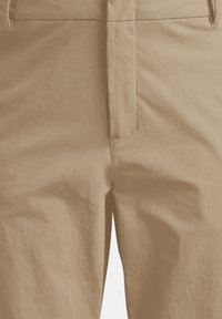 Gros plan sur le devant d'un pantalon beige avec des passants de ceinture visibles, des coutures apparentes et des détails de la fermeture éclair.