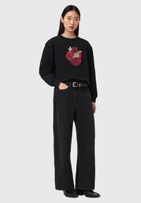 Νεαρή γυναίκα με μακριά μαύρα μαλλιά φοράει μαύρο φούτερ με σχέδιο ανατομικής καρδιάς σε κόκκινο, μαύρο τζιν με φαρδιά μπατζάκια, ζώνη και μαύρα μπότες.