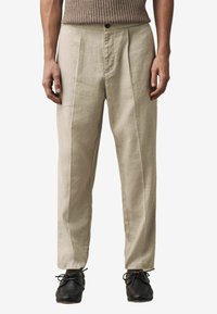 Pantalons en lin beige avec une coupe décontractée, dotés de plis avant, d'une fermeture à bouton unique, portés avec des chaussures en cuir noires.