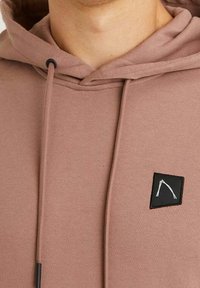 Růžová mikina s kapucí a šňůrkou, na hrudi s černým čtvercovým logem. Materiál má hladkou strukturu a uvolněný design.