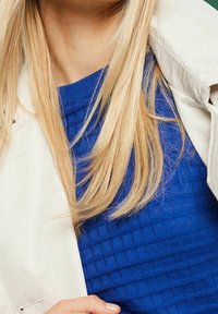 Femme blonde portant un pull bleu texturé et une veste beige clair drapée sur son épaule, tenant la veste d'une main.