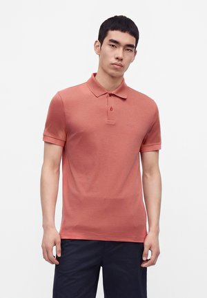 Jeune homme portant un polo manches courtes de couleur saumon et un pantalon foncé, debout devant un fond blanc uni.