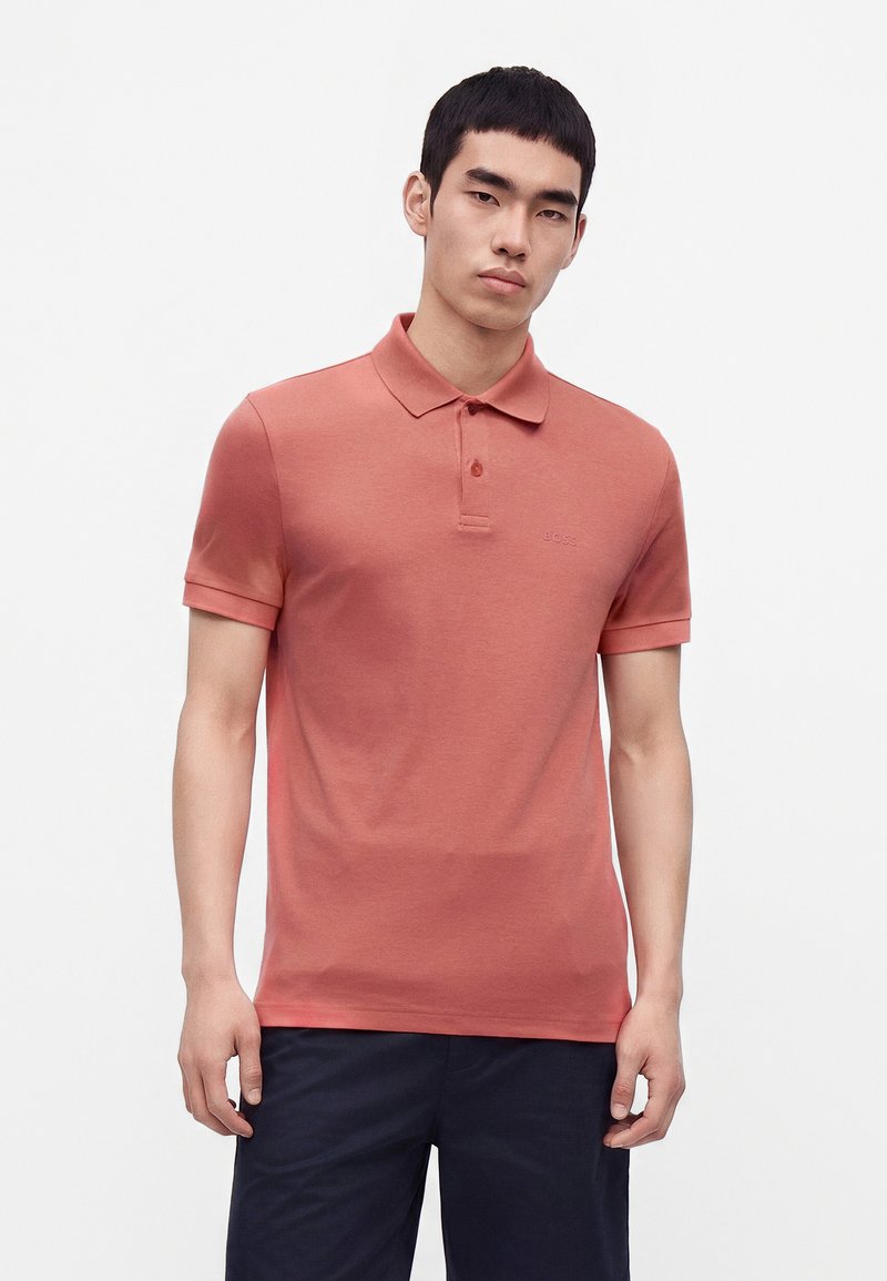 Jeune homme portant un polo manches courtes de couleur saumon et un pantalon foncé, debout devant un fond blanc uni.