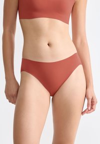 Donna che indossa un set di intimo terracotta senza cuciture, mostrando la parte centrale e la parte superiore delle gambe su uno sfondo neutro.
