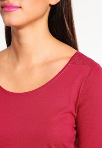 Mörkröd långärmad topp med vid nacklinje och spetsdetaljer på axlarna. Slät textur, lättviktsmaterial.