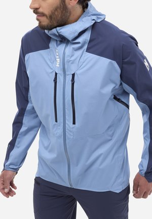 Mann trägt eine hellblaue und marineblaue Kapuzenjacke mit vorne und seitlich angebrachten Reißverschlusstaschen, steht mit leicht nach vorn gehaltenen Händen.