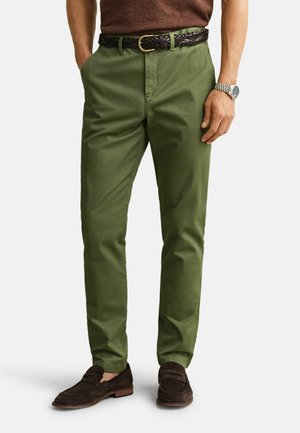 Homme portant un pantalon chino vert olive avec une ceinture tressée marron, des mocassins marron, une chemise marron et une montre-bracelet argentée au poignet gauche.