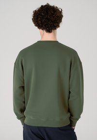 Asmuo su garbanotais plaukais, dėvintis paprastą alyvuogių žalią megztinį ir tamsias kelnes, stovi nugara į neutralią foną.