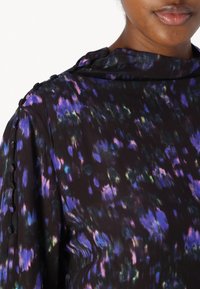 Long-sleeve blouse in dark fabric featuring a colorful floral pattern with shades of purple and green; includes button accents on the shoulder.