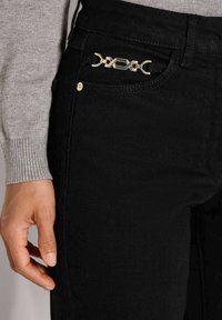 Nahaufnahme einer Hand neben einer schwarzen Hose mit einer kleinen goldenen dekorativen Kette an der vorderen Tasche und einem grauen Langarmoberteil.