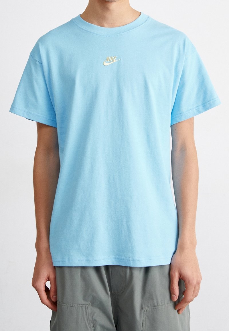 Blå bomullströja med korta ärmar, rund halsringning och en liten gul Nike-logotyp broderad på bröstet. Slät textur.