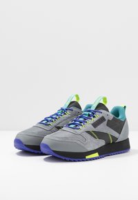 Šedé sportovní boty z mesh a semišových materiálů. Mají jasně modré a zelené šňůrky, černou podrážku a výrazné cik-cak prošití.