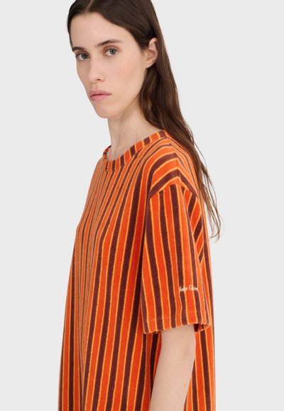 Žena s dugom smeđom kosom nosi preveliku košulju s narančastim i smeđim okomitim prugama, stoji na jednostavnoj svijetlosivoj pozadini.