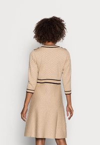 Beige stickad klänning med ett strukturerat mönster, 3/4-långa ärmar, svarta detaljer och gyllene knappdetaljer på axlarna. A-linjär siluett.