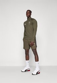 Olijfgroene sportjack met rits en logo, bijpassende shorts, witte sneakers met rode accenten en knielange witte sokken.