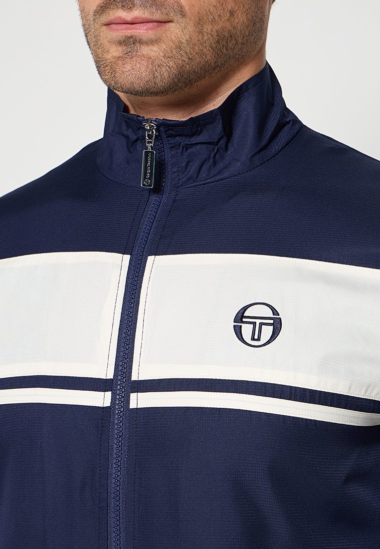Giacca a track navy con strisce orizzontali bianche, colletto alto e zip frontale. Presenta un piccolo logo circolare sul lato sinistro del petto.