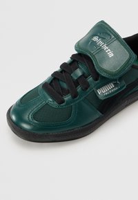 Baskets Puma vert foncé et noir avec des lacets noirs, "Spökerin" sur la languette et une plaque métallique "Puma Palermo" sur le côté.