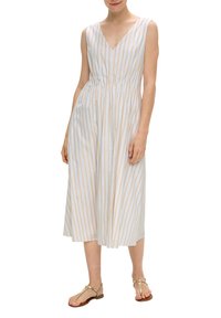 Robe blanche avec des rayures verticales dorées et beiges, décolleté en V, sans manches, taille cintrée, jupe fluide, portée avec des sandales beiges.