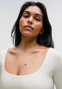 Una persona con cabello largo y oscuro lleva puesto un prenda ajustada color crema con un escote cuadrado, que muestra una notable marca de belleza en el pecho.