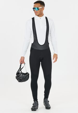 Mies pukeutuneena mustiin pyöräilyhousuihin, valkoinen pitkähihaiseen pelipaitaan, heijastavat aurinkolasit, oikeassa kädessä musta pyöräilykypärä.