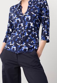 Camisa abotonada en azul marino con patrones abstractos en azul y blanco, con mangas tres cuartos, cuello clásico y textura suave.