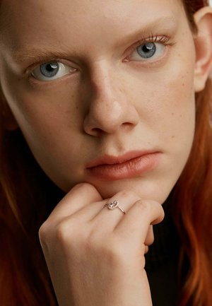 Anillo de plata con un engaste ovalado que presenta una gema central, llevando en una mano que descansa contra la cara, con cabello rojo de fondo.