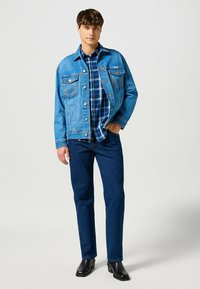 Jeune homme portant une veste en denim bleu, une chemise à carreaux bleue, un jean bleu foncé et des bottes en cuir noires, se tenant contre un fond uni.