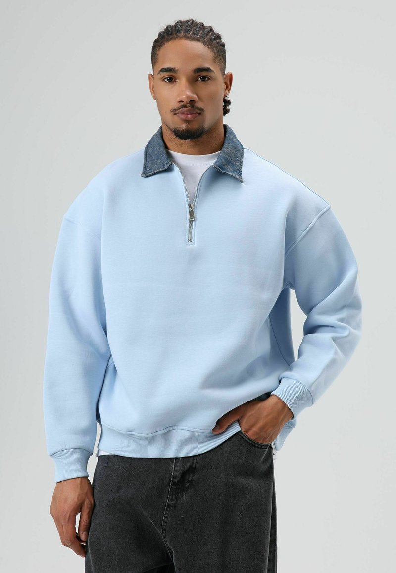 Sweat-shirt bleu clair avec col zippé et accents en denim. Présente des épaules tombantes et des poignets côtelés. Porté avec un pantalon gris foncé.