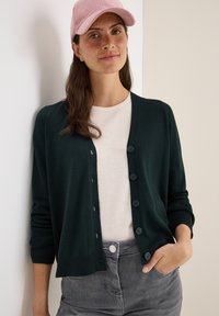 Donkergroene cardigan met een knoop-voorontwerp, geribbelde manchetten en een lichtbeige t-shirt eronder. Model draagt een lichtroze pet.