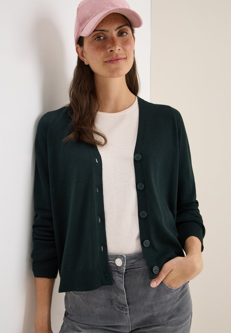 Donkergroene cardigan met een knoop-voorontwerp, geribbelde manchetten en een lichtbeige t-shirt eronder. Model draagt een lichtroze pet.