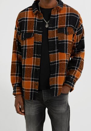 Mand iført orange og sort ternet skjorte med knapper over en sort T-shirt og falmede sorte jeans, med den ene hånd i lommen.
