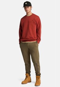 Röd tröja, olivgröna sweatpants och tanfärgade stövlar. Byxorna har en Timberland-logotyp tryckt längs benet. Svart keps med logotyp.