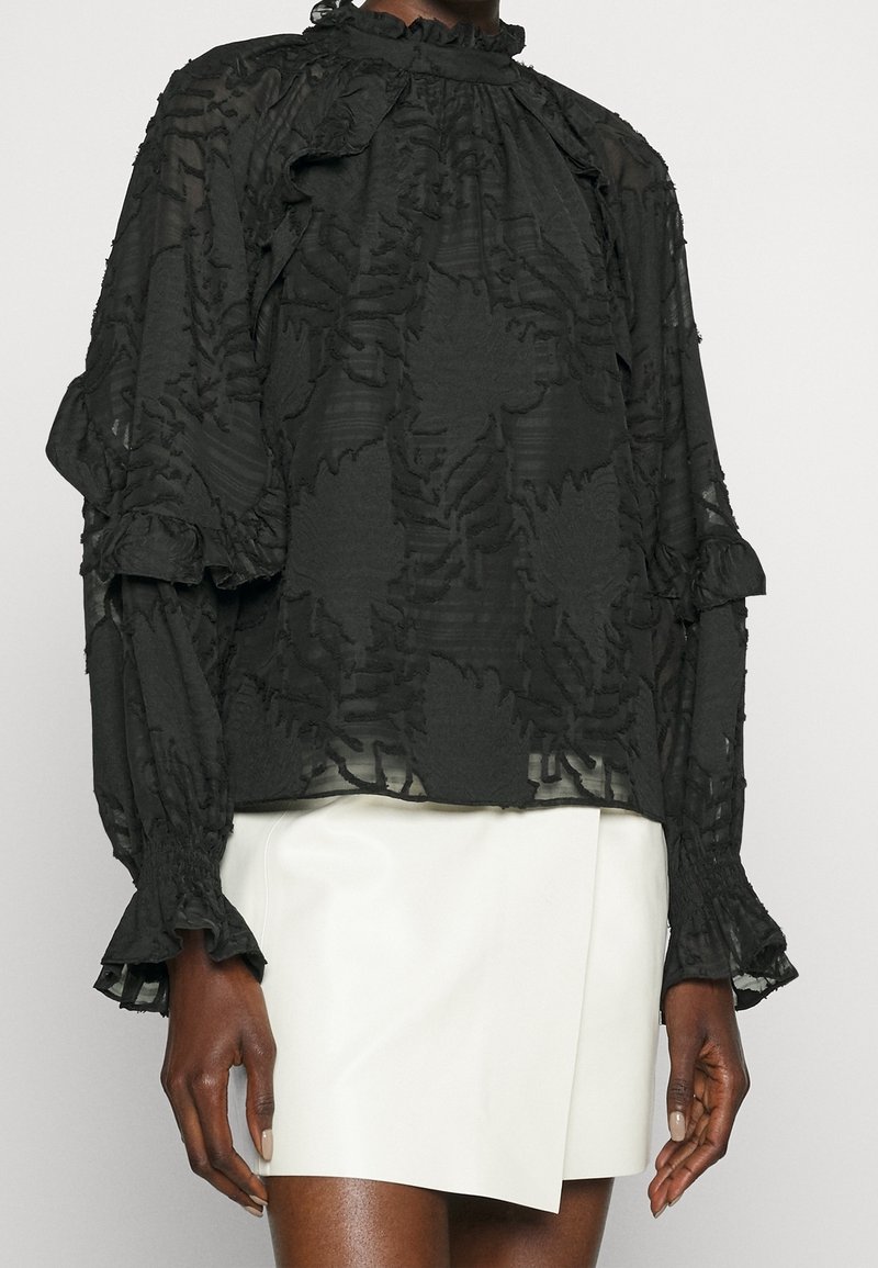 Blouse noire transparente avec motif floral texturé et poignets à volants, associée à une mini-jupe blanche asymétrique sur une personne debout.