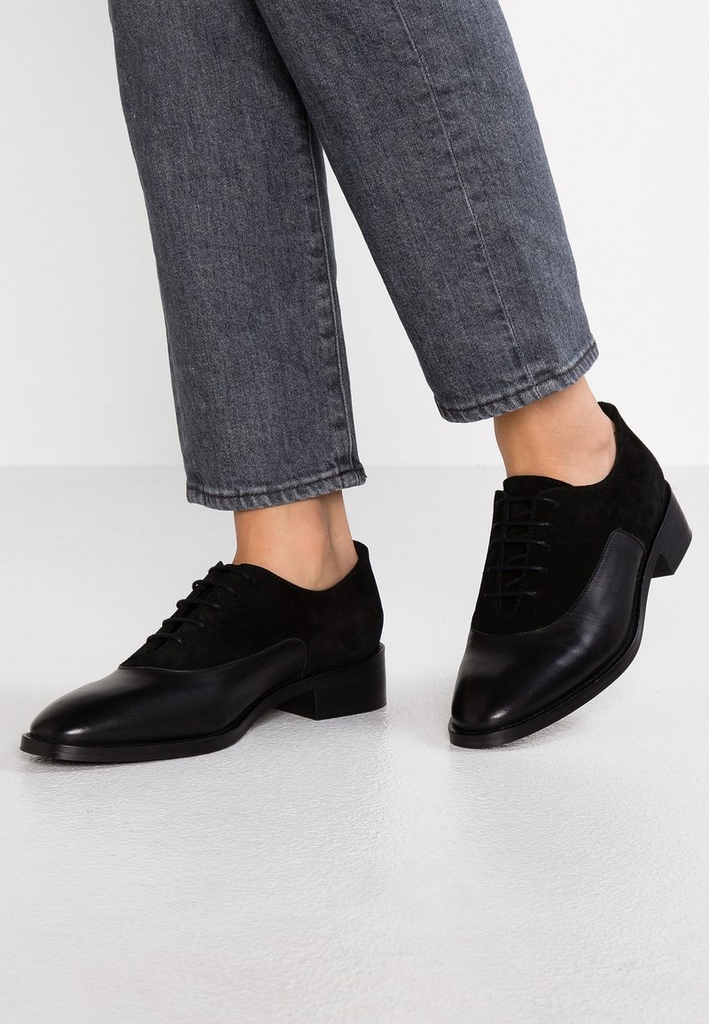 Chaussures Oxford en cuir et suède noir portées avec un jean gris foncé longueur cheville sur un sol clair avec un fond blanc.