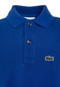 Niebieski polo z teksturą, z dwiema ciemnymi guzikami i małym zielonym logo krokodyla na prawej części klatki piersiowej.