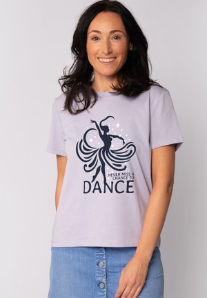 Vrouw met donker haar die een lila shirt draagt met een dansende silhouet en de tekst "Never miss a chance to dance" en een blauw rokje met knopen.