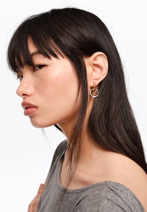 Joven con cabello largo y oscuro, lleva pendientes de aro dorados y plateados y una camiseta gris, mirando de lado contra un fondo blanco.