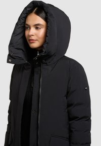 Sort polstret jakke med stor, justerbar hætte, lynlås foran, trykknapper og sidelommer. Glat tekstur med diskret mærkedetalje.