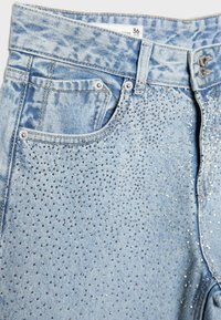 Hellblaue Jeans aus Denim mit kleinen metallischen Nieten, die dicht die Vordertasche und den oberen Oberschenkelbereich bedecken, ausgestattet mit einem Doppelknopfverschluss und Gürtelschlaufen.