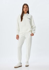 Weißer Pullover mit hohem Kragen und subtiler Markenkennzeichnung, kombiniert mit passenden Jogginghosen mit Seitentaschen und schmal zulaufenden Bündchen.