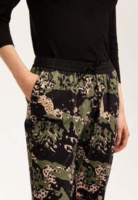 Pantalones jogger de camuflaje con cintura elástica y cordón, principalmente en verde y negro con acentos en beige y un ajuste relajado.
