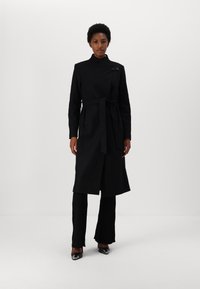 Cappotto lungo nero con cintura e colletto alto, realizzato in tessuto liscio, con un fronte asimmetrico e una chiusura decorativa nera.