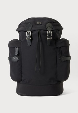 Sac à dos noir avec deux boucles à l'avant, des poches latérales et un rabat supérieur, présenté sur un fond uni clair.