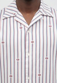 Κοντινό πλάνο λευκού κοντομάνικου πουκαμίσου με κατακόρυφες κόκκινες και μπλε ρίγες και μικρά κόκκινα διαμαντένια λογότυπα, που φοριέται από ένα άτομο.