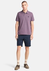 Fialová polo tričko s malým černým logem, spárováno s námořnickými šortkami. Model má na sobě hnědé boty a krémové ponožky s hnědými pruhy.