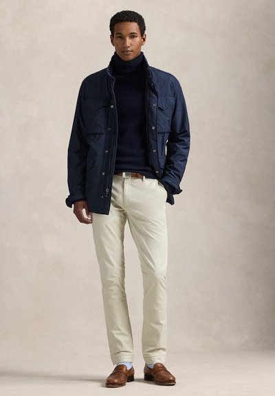 Muž v tmavomodrej bunde cez čierny rolák, béžové nohavice a hnedé mokasíny stojí na neutrálom pozadí.